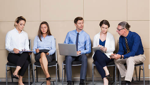 Five male and female business people sitting in row at wall and waiting for job interview. They are working on laptop, using smartphone and writing.