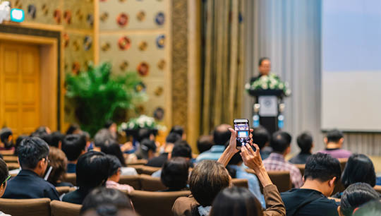 Back side of audience taking photo by cellphone the Speaker with podium and presentation